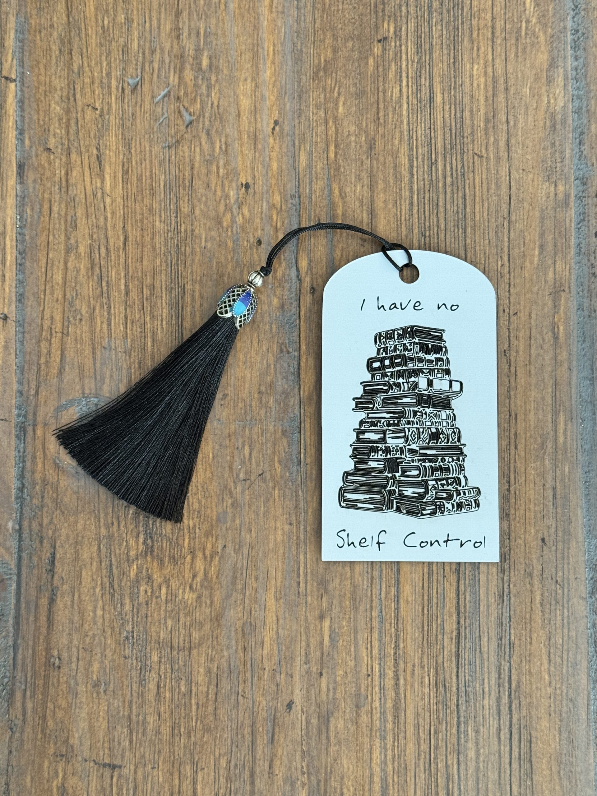 White acrylic bookmark with an engraved quote "I have no shelf control", an illustration of an unruly stack of books, and a black tassel.